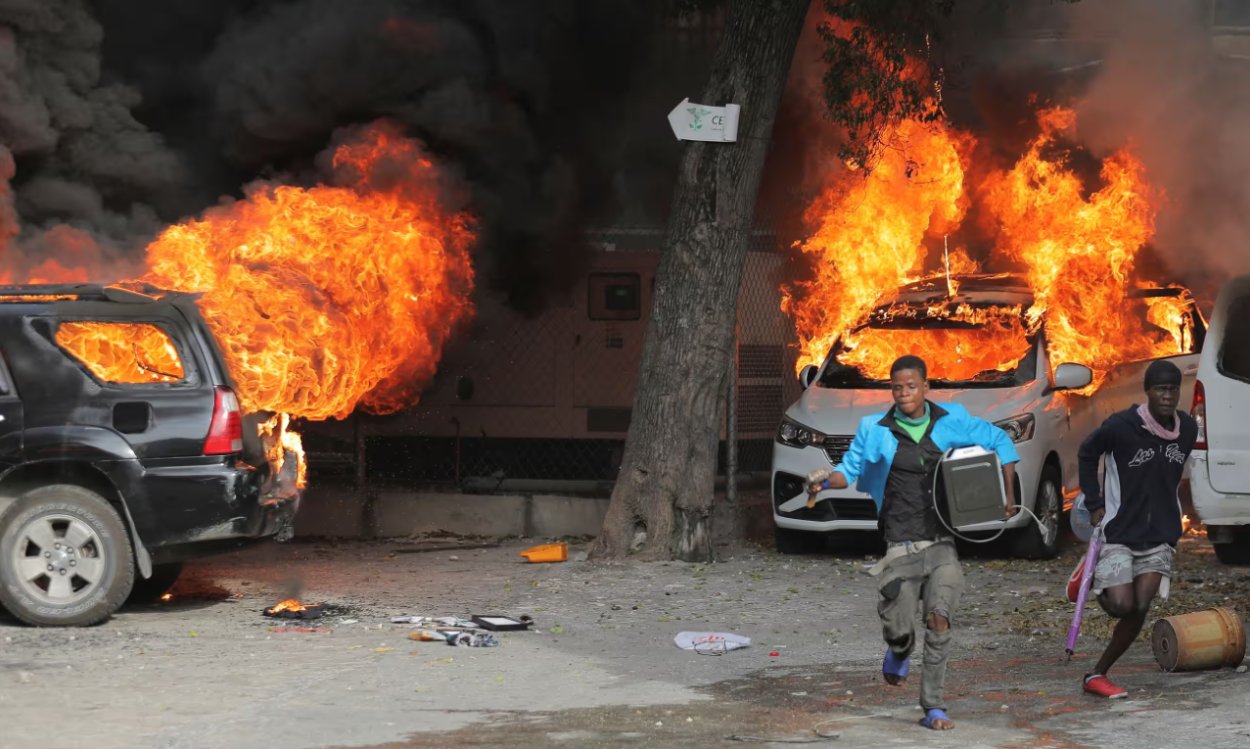 Muži behajú s ulúpeným tovarom v blízkosti horiacich áut počas minuloročných protestoch proti rastúcim cenám pohonných hmôt a kriminalite v Port-au-Prince.